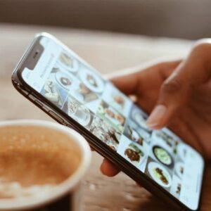 Foto en tonalidades sepia de una mano sujetando un móvil donde se puede ver el instagram con fotos de comida, al lado, junto a sus manos hay un café.