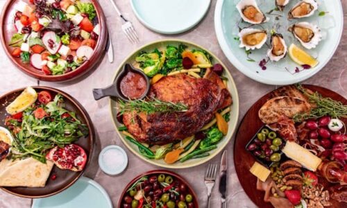 Seis platos con grandes cantidades de comida en una mesa. Se puede ver carne, ensalada, mejillones, aceitunas y patatas fritas.