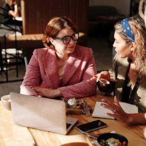 Dos chicas mujeres hablando en frente de una mesa donde hay un ordenador y papeles.